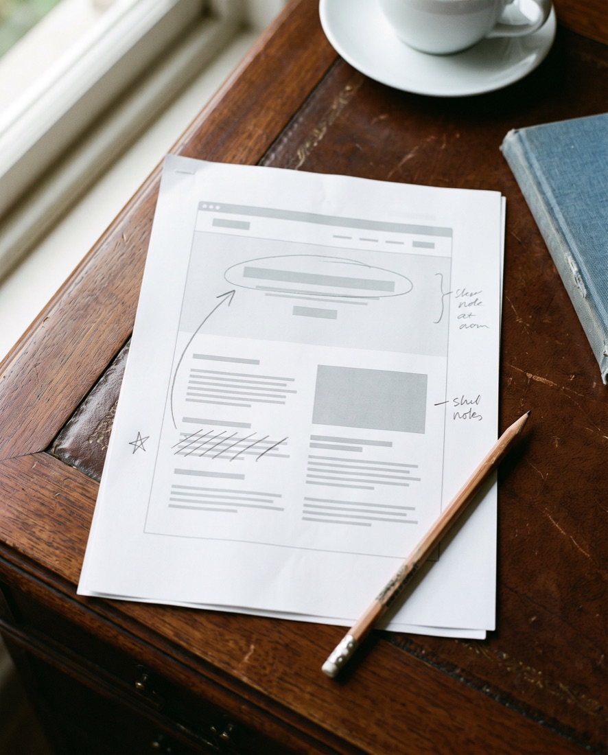 A printed website page on a desk, reviewed by hand in pencil: the headline circled, an arrow moving a paragraph up, one block crossed out and notes in the margin.